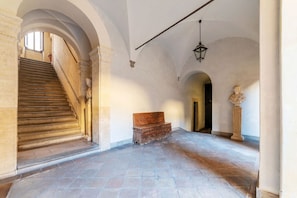 Eine prachtvolle Treppe führt zu einer oberen Ebene, an der ein Sitzplatz und eine Statue in einem geräumigen, mit Bögen versehenen Gang platziert sind.
