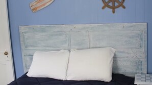 Een bed met een blauw hoofdeinde, witte kussens en een houten stuurwiel aan de muur.