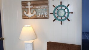 Een kamer met een witte muur, een houten ankerschild, een scheepsschroef, een lamp, een klein wit kastje en een bruine stoel.