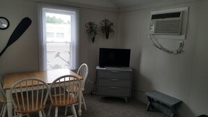 Een kleine kamer met een houten tafel en stoelen, een televisie op een standaard, een wandairconditioner en een raam met jaloezieën.