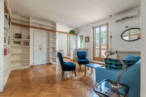 Ein Wohnzimmer mit Holzboden, einem blauen Sofa, einem blauen Sessel, einem Tisch, einer Lampe, einem Bücherregal und einer Heizung.