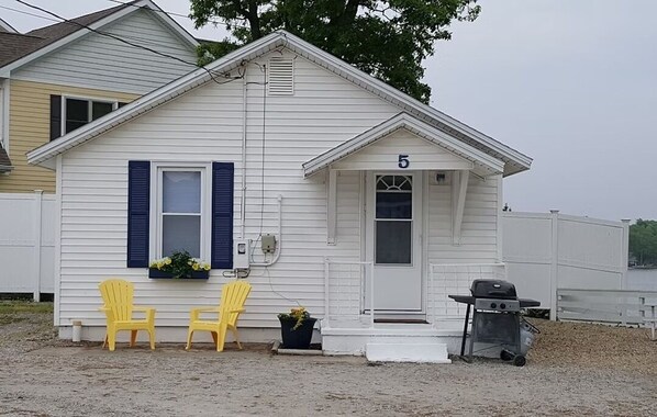 A single-story house with a white exterior, number 5, featuring two yellow chairs outside, a small porch, and a grill. - Cabin 5 at Dockside Lake Resort (Monticello)