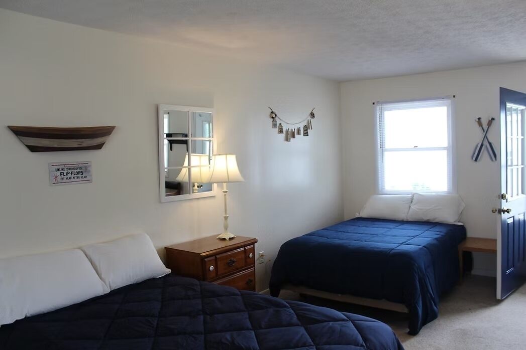 A bedroom with two beds, a nightstand, a lamp, a mirror, a window, and a door.
