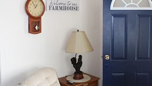 Ett rum med en vit fåtölj, ett träbord vid sängen med en lampa, en väggklocka och en dörr med en skylt som lyder: ”Välkommen till vårt GÅRDSHUS”.