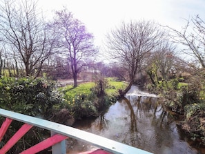 A river with trees and greenery, a bridge with red and white railings, and a grassy area in the background. - Fullers Studio (Holyhead)