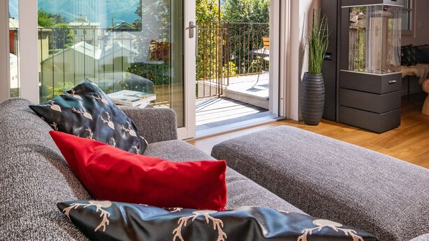 A modern living room with a grey sofa, red and black patterned cushions, a large window with a mountain view, and a wood-burning stove. - Stella - panoramic attic in the BOTTONERA casa vacanza in alpine design (Chiavenna)