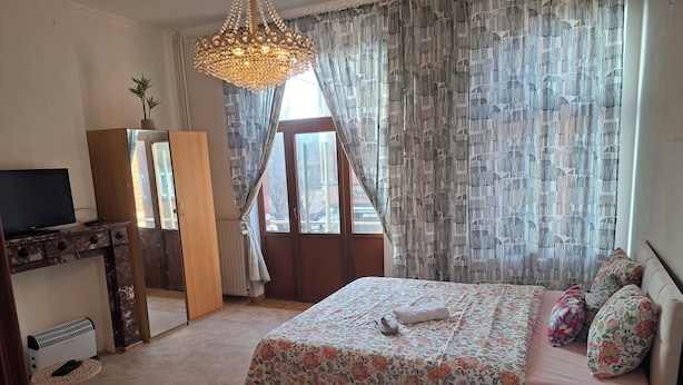 Un dormitorio con cama, televisor sobre un mueble, radiador, ventana con cortinas y un candelabro.
