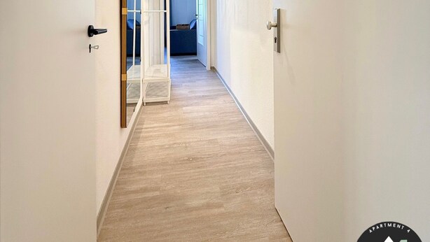 Un couloir avec un plancher en bois, des murs blancs et un ventilateur de plafond. Une porte avec un miroir et une petite marche sont visibles sur la gauche.