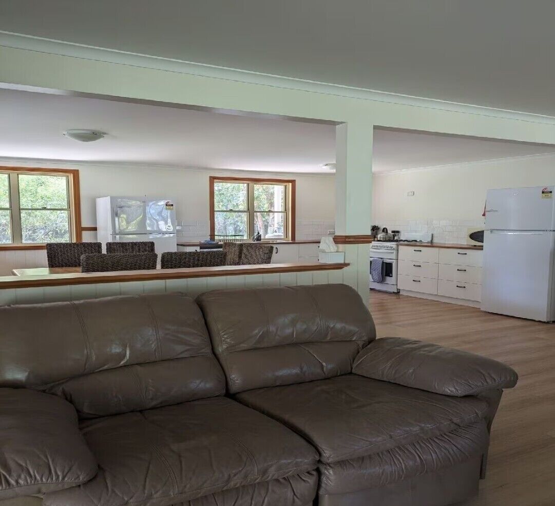 A living room with a brown leather sofa, a kitchen with white cabinets and a refrigerator, and large windows with a view of greenery.