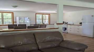 A living room with a brown leather sofa, a kitchen with white cabinets and a refrigerator, and large windows with a view of greenery.