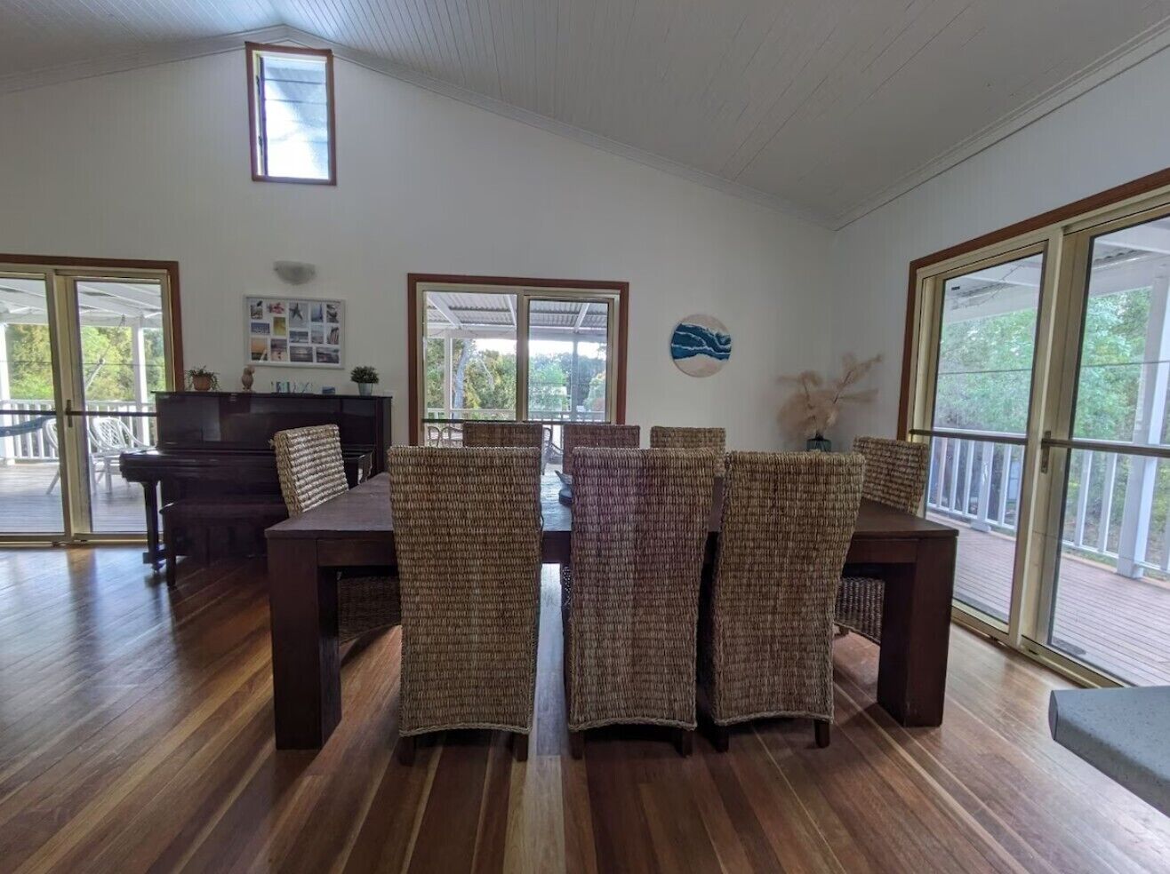 A spacious dining area with a wooden table and wicker chairs, large windows, and a piano.