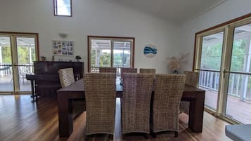 A spacious dining area with a wooden table and wicker chairs, large windows, and a piano.