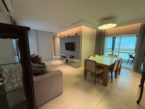 A modern living room with a sofa, a glass coffee table, a dining table with chairs, a flat-screen TV mounted on a wooden wall, and a large window with curtains. - Enjoy a Convenient Location and Comfort in Jardim Goias - Mfes (Goiânia)