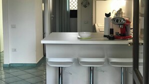 A kitchen with a white countertop, a coffee machine, and a window with a patterned blind. - Exclusive Villa on the Pelosa Beach (Stintino)