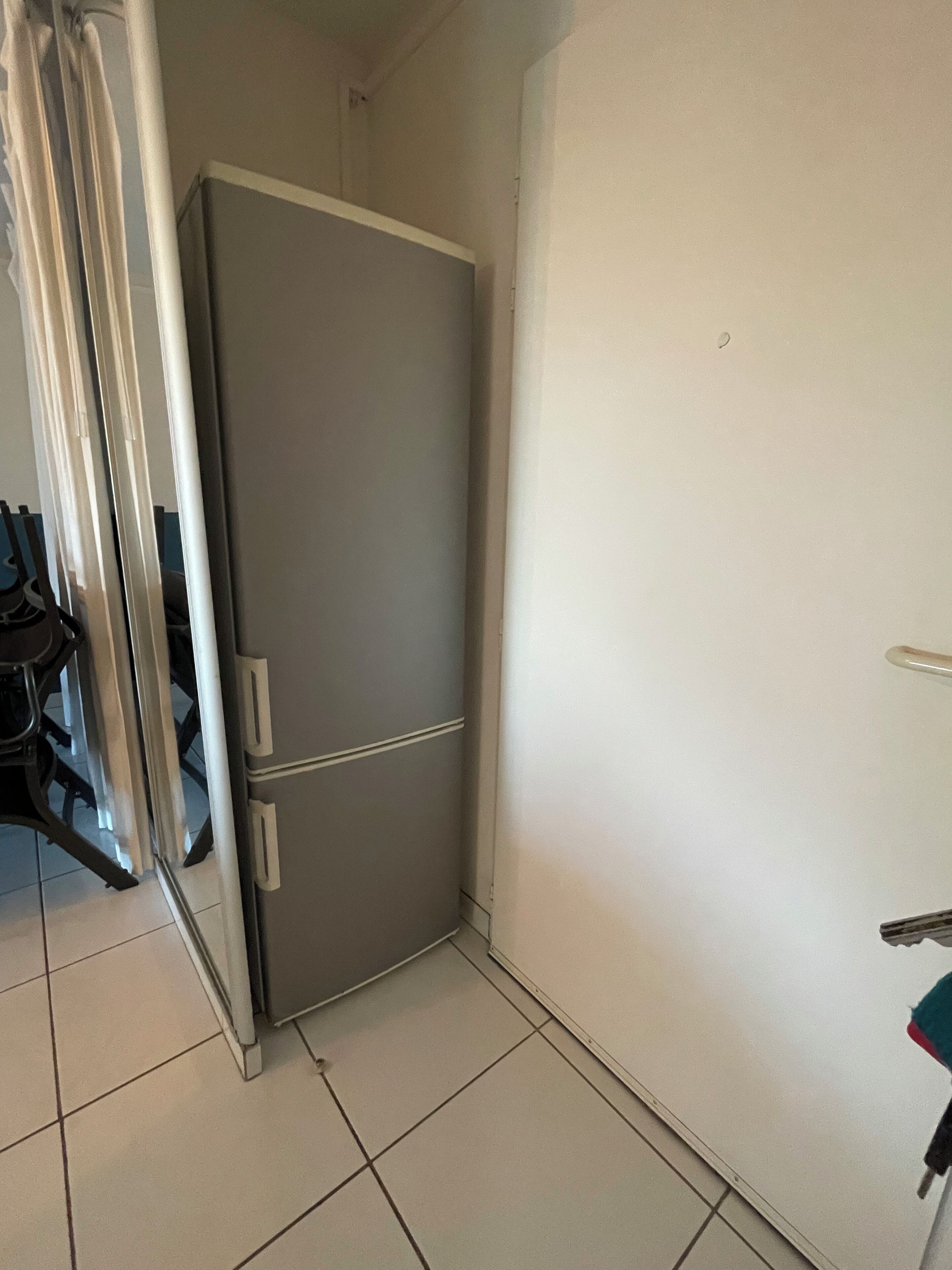 Een kleine keuken met een grijze koelkast, witte kastjes en een betegelde vloer.