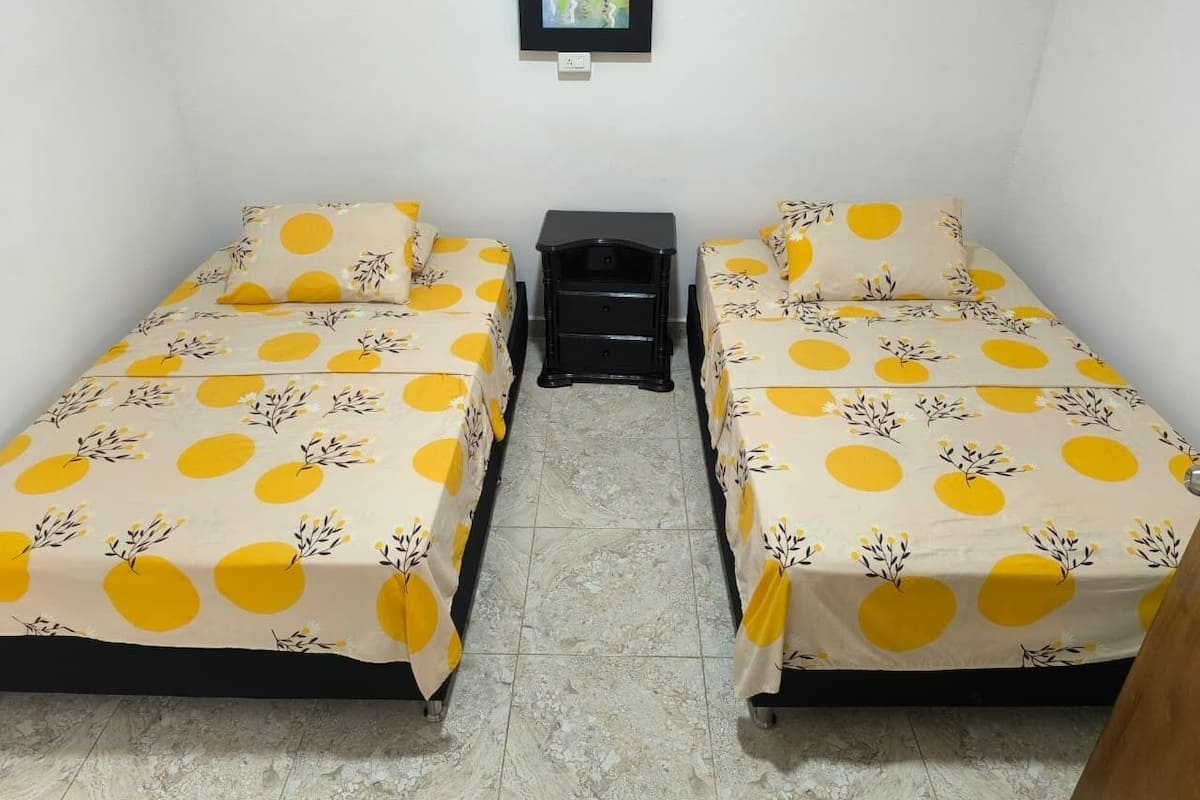 Une chambre avec deux lits, chacun recouvert d’une couvre-lit à pois jaunes et blancs, une table de chevet noire et une petite table sur laquelle est posé un vase de fleurs.