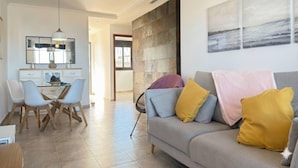 Una sala de estar moderna con un sofá gris, cojines amarillos y una zona de comedor con sillas blancas y una mesa.
