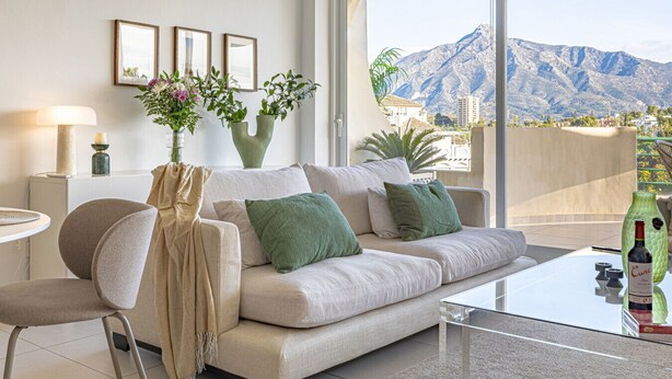 Un salón moderno con un sofá, cojines verdes, una mesa de centro de vidrio y vistas a montañas.