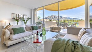 Una sala de estar moderna con sofá, mesa de centro y sillones, una mesa de vidrio con una botella, y un ventanal con vista a la montaña.