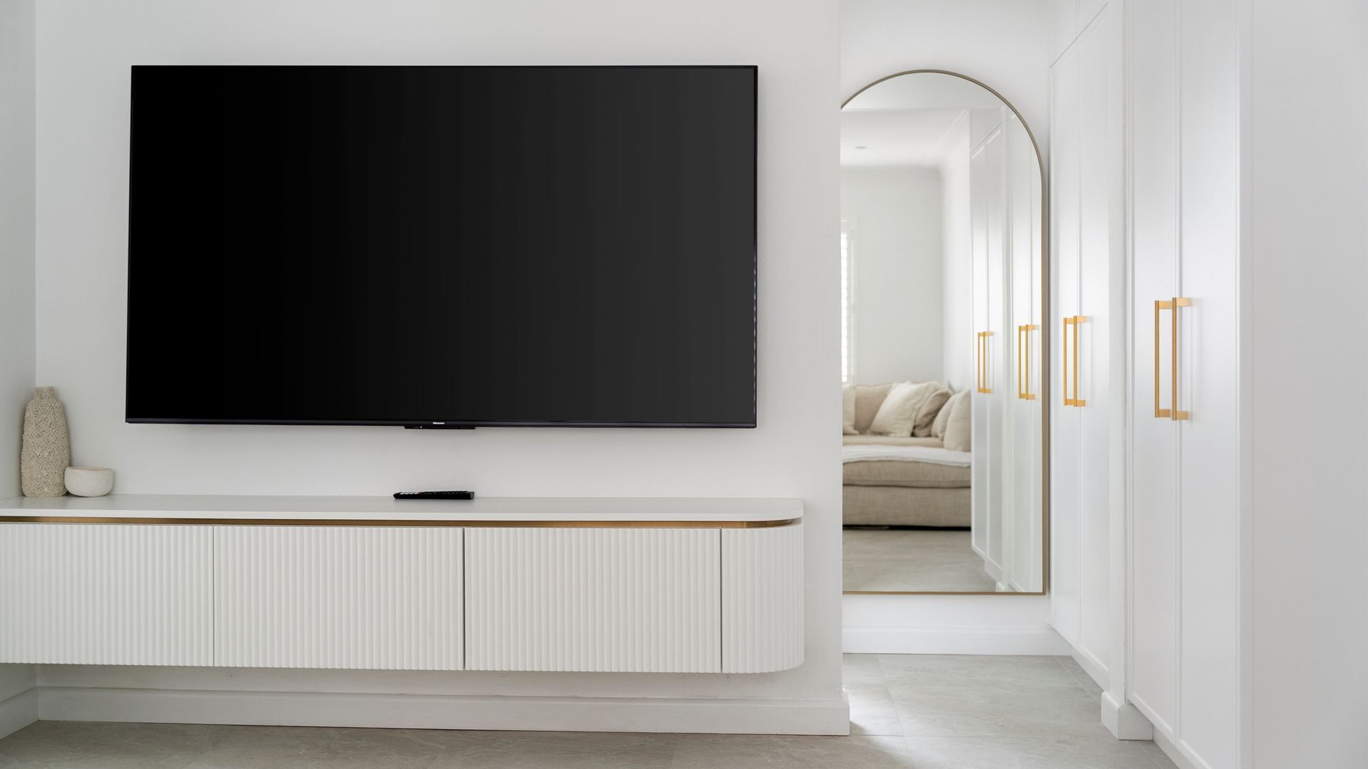 A modern living room with a large flat-screen TV mounted on a white wall, a minimalist white console table below, and a doorway leading to another room with a sofa and a mirror.