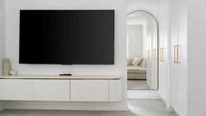Un saló modern amb un gran televisor de pantalla plana muntat sobre una paret blanca, una taula baixa blanca minimalista a sota i una porta que condueix a una altra habitació amb un sofà i un mirall.