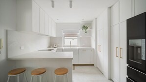 Una cuina moderna amb armaris blancs, una illa central amb tamborets, forn integrat i nevera.
