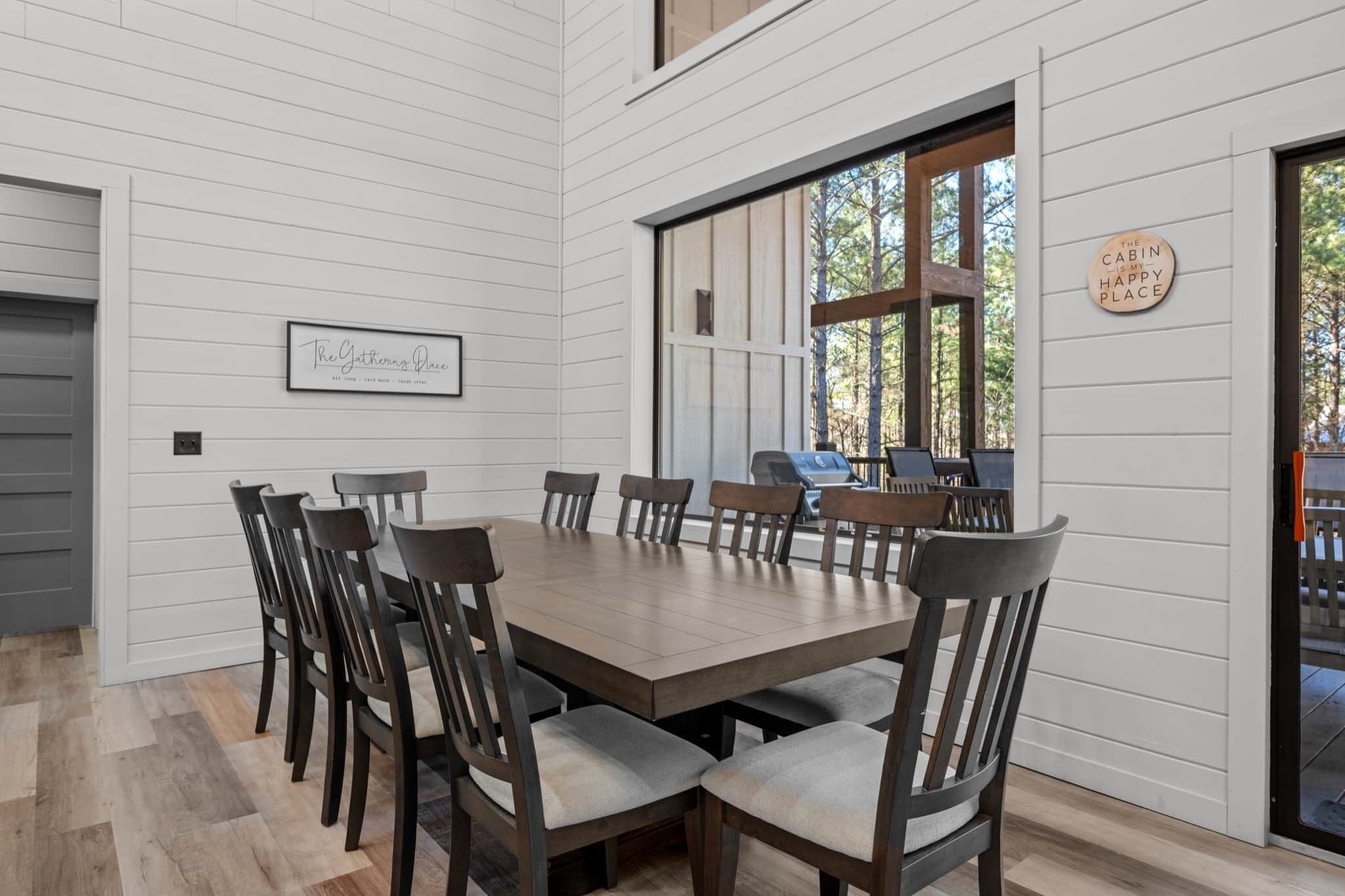 Un comedor con una gran mesa de madera y sillas, un cartel enmarcado en la pared, y una ventana con vista a los árboles.