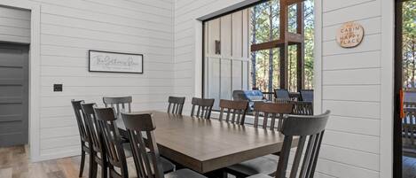 Un comedor con una gran mesa de madera y sillas, un cartel enmarcado en la pared, y una ventana con vista a los árboles.
