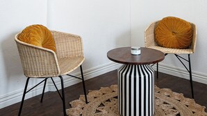 Una habitación con dos sillas de mimbre con cojines amarillos, una mesa redonda con base a rayas blancas y negras, y un tapiz tejido. Un cuadro enmarcado de una escena de playa cuelga en la pared.