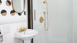 Un baño con lavamanos blanco, detalles dorados, un espejo y una cabina de ducha de vidrio.