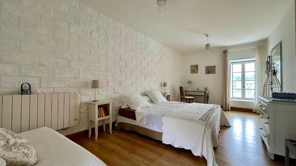 A bedroom with a white brick wall, a bed with white bedding, a bedside table with a lamp, a chair, and a dresser.