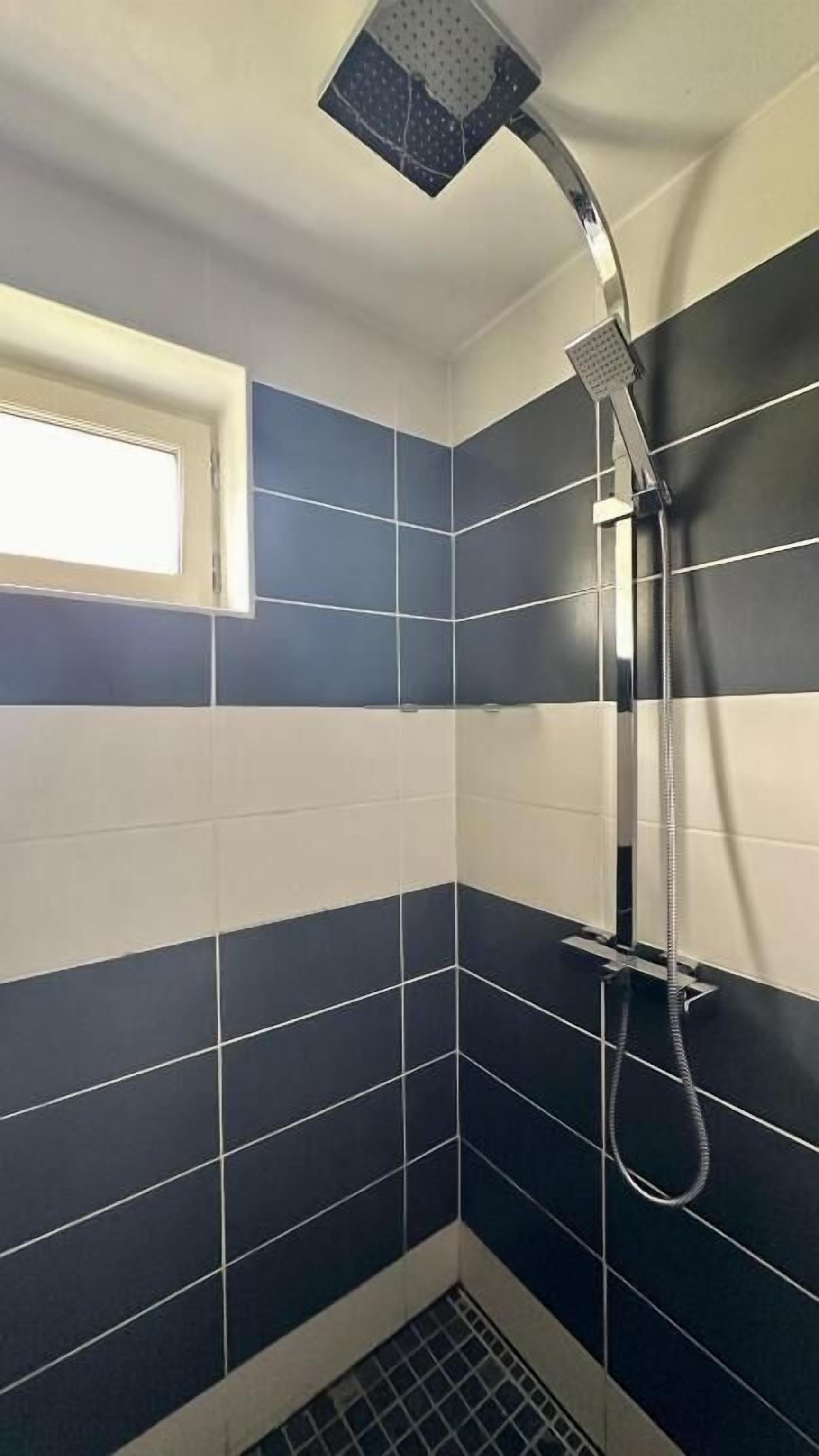 Ein kleines Badezimmer mit dunkelblauen und weißen Fliesen, einer Dusche mit Handbrause und einem Fenster.