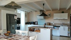 A modern kitchen with white cabinetry, a central island, and a dining area with a table and chairs. - Clos Beldane (Le Tâtre)