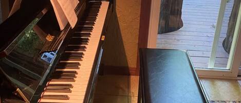 A piano with sheet music, a bench, and a view of a wooden deck and greenery outside.