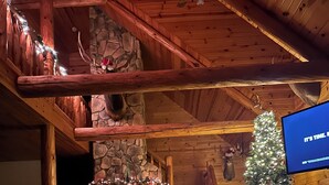 A cozy log cabin interior with a stone fireplace, a Christmas tree, and a television screen displaying "It's time." - Mountain feel/10 minutes from the city! (Pittsburgh)