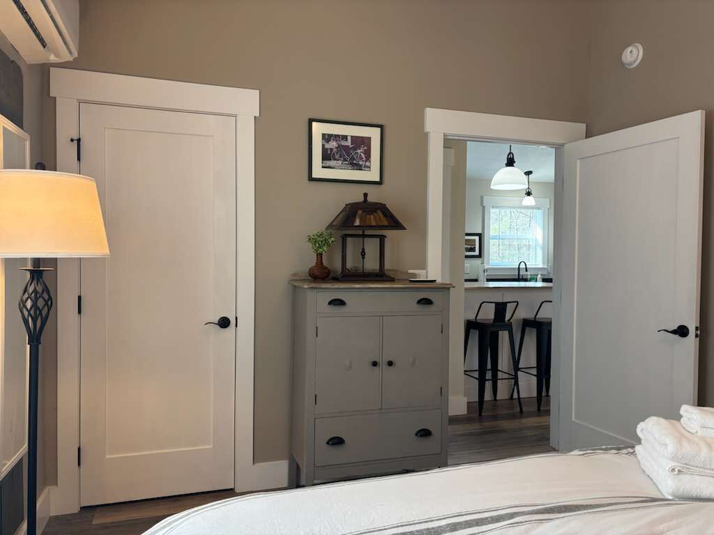A bedroom with a bed, a dresser, a lamp, a picture on the wall, and a door leading to a bathroom.
