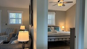 Quarto de hotel com cama, ventoinha no teto, mesa de cabeceira com abajur, sofá e janela com cortinas.
