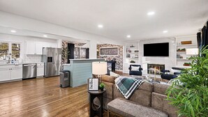 Ein geräumiges Wohnzimmer mit einer Couch, einem Couchtisch und einem Fernseher. Es gibt eine Küche mit weißen Schränken und Edelstahlgeräten.
