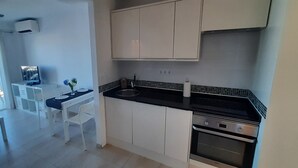 Cozinha moderna com armários brancos, bancada preta e forno embutido. Possui área de jantar com mesa e cadeiras brancas, e uma varanda com vista para a água.