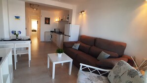 Sala de estar com sofá marrom, mesa de centro branca e televisão. Há também uma área de cozinha com frigorífico e micro-ondas.
