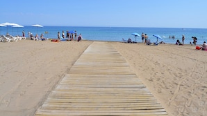 Um calçadão de madeira que leva a uma praia arenosa, com guarda-sóis e espreguiçadeiras.