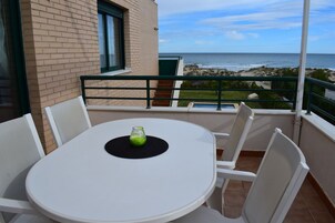 Un balcón con mesa y sillas blancas, un vaso de agua y vista a la playa y al océano.