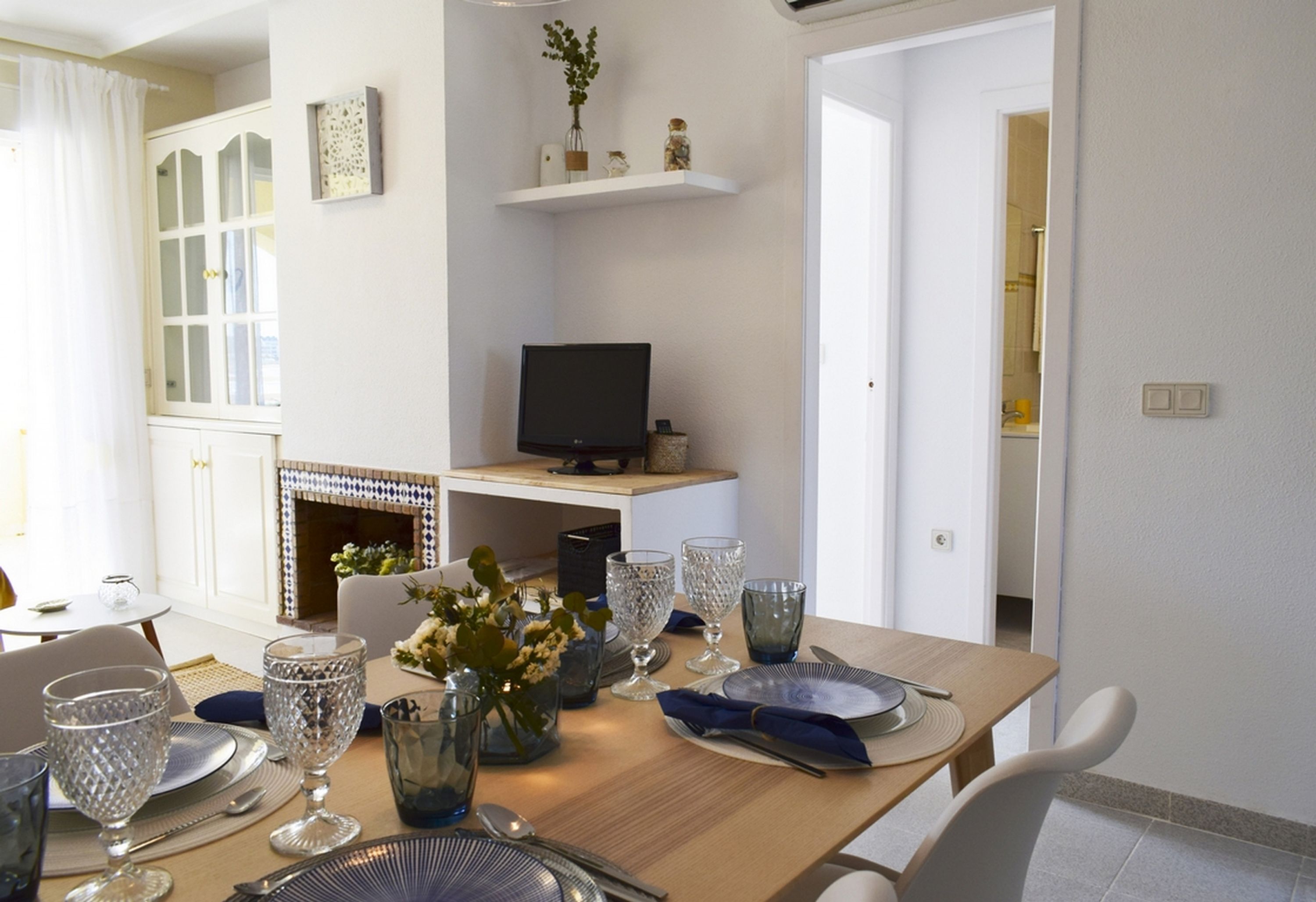Un coin repas avec une table en bois dressée pour quatre personnes, entourée de chaises. On aperçoit un téléviseur posé sur un meuble au-dessus de la table, et une cheminée est visible au fond.
