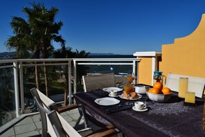 Mesa de comedor lista con platos, cubiertos y un cuenco de naranjas en un balcón con vista al mar.