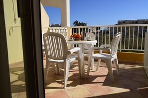 Ein Balkon mit weißen Plastikstühlen und einem Tisch, einem Korb mit Orangen und einer Kaffeekanne.