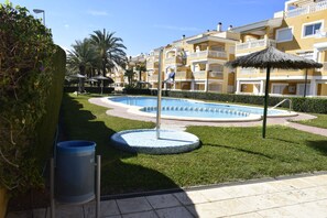 Ein Swimmingpool, umgeben von einer gepflegten Rasenfläche, mit einem Reetdachschirm und einem Mülleimer in der Nähe.