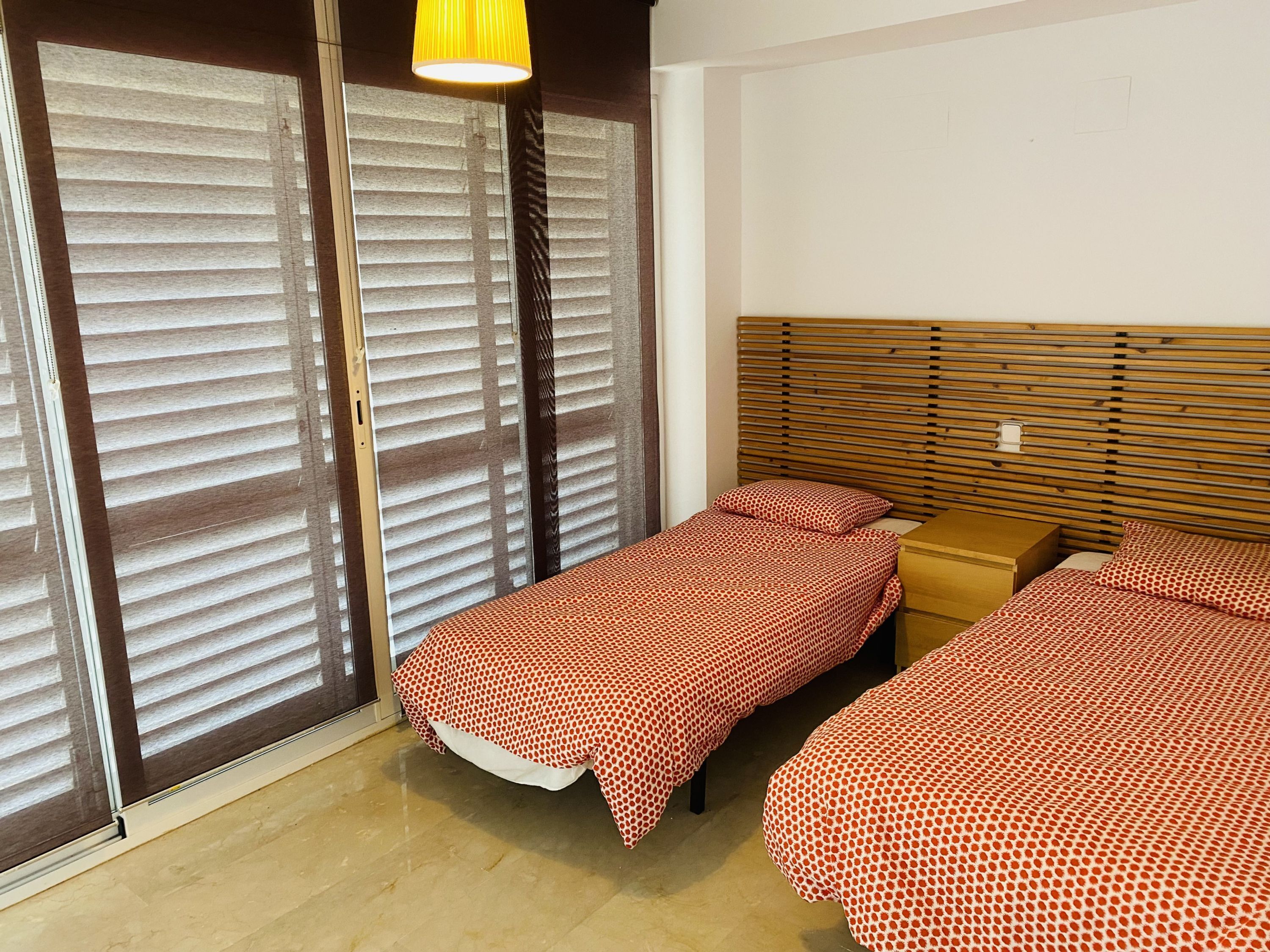 A room with two beds, a wooden headboard, a nightstand, and a window with blinds.