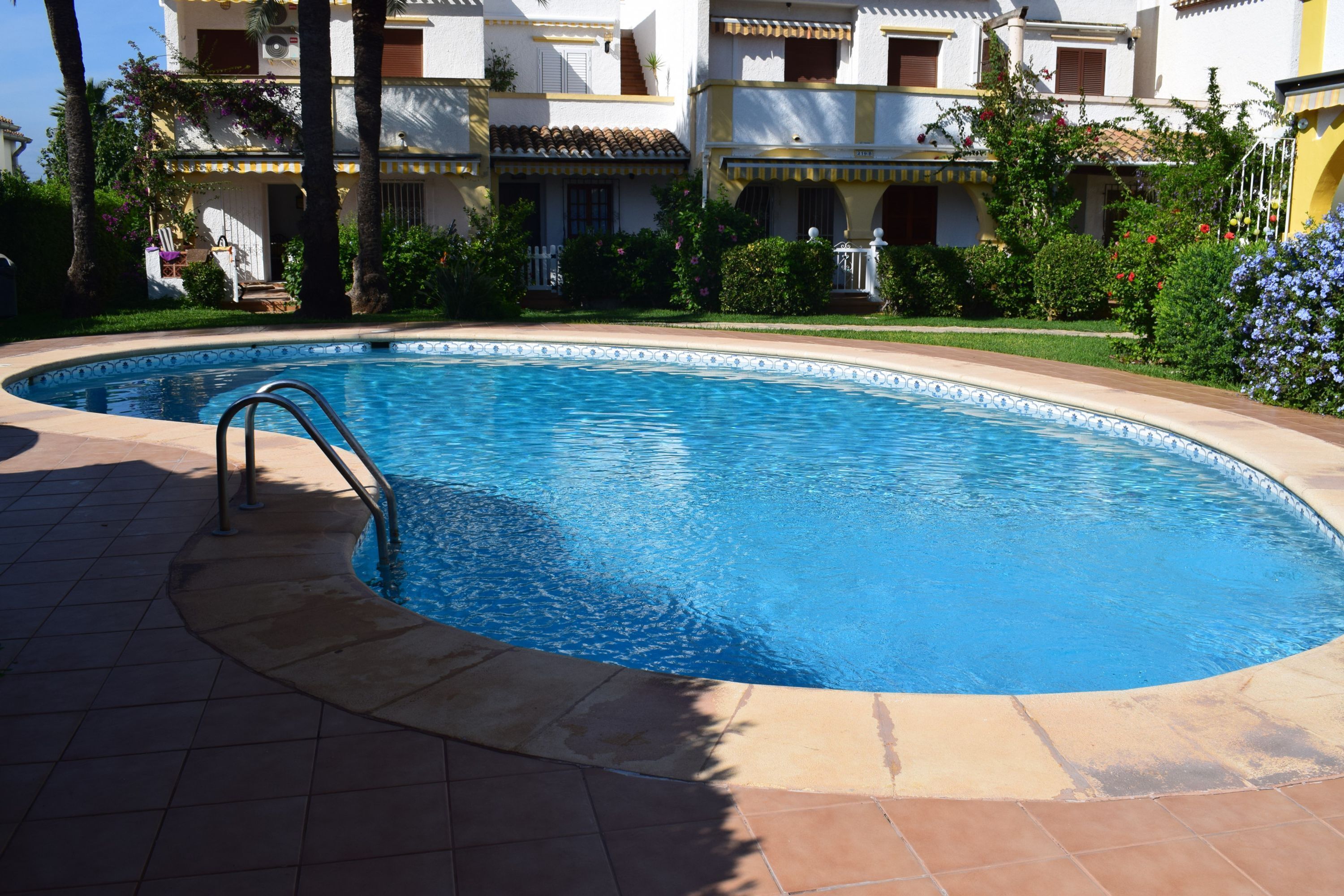 A swimming pool with a tiled deck, surrounded by a well-maintained garden and a multi-story building with balconies.