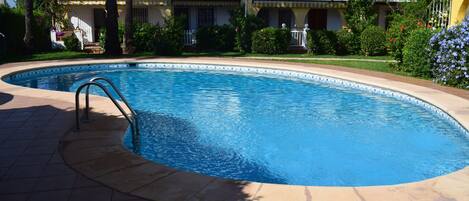 A swimming pool with a tiled deck, surrounded by a well-maintained garden and a multi-story building with balconies.