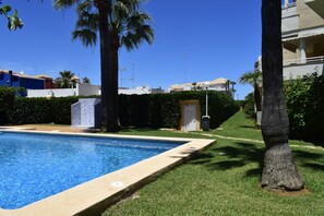 A swimming pool surrounded by well-maintained grass and palm trees, with buildings in the background. - Ap2236 in Denia With 2 Bedrooms and 2 Bathrooms (Denia)